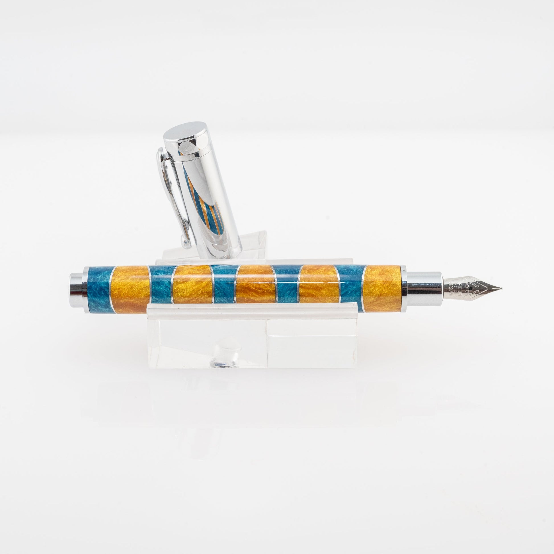 A handmade blue and gold resin and aluminum fountain pen rests on a clear stand. The plating is chrome and it features a German Iridium nib.