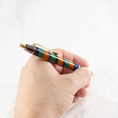 Orange, purple, and green handmade striped resin ballpoint click pen with gold and gunmetal plating being held in a hand.