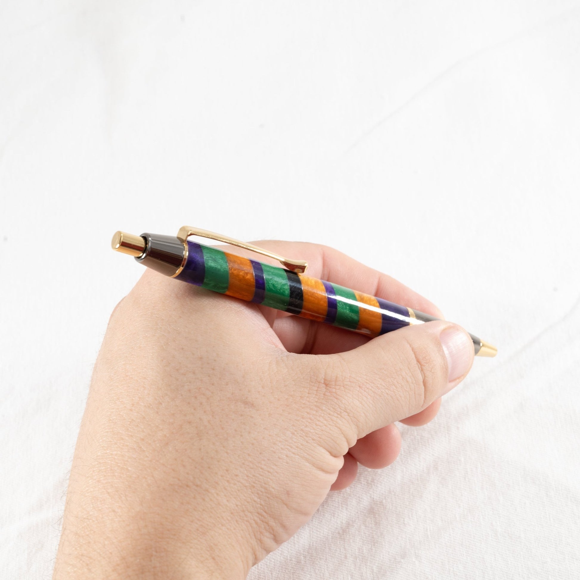 Orange, purple, and green handmade striped resin ballpoint click pen with gold and gunmetal plating being held in a hand.