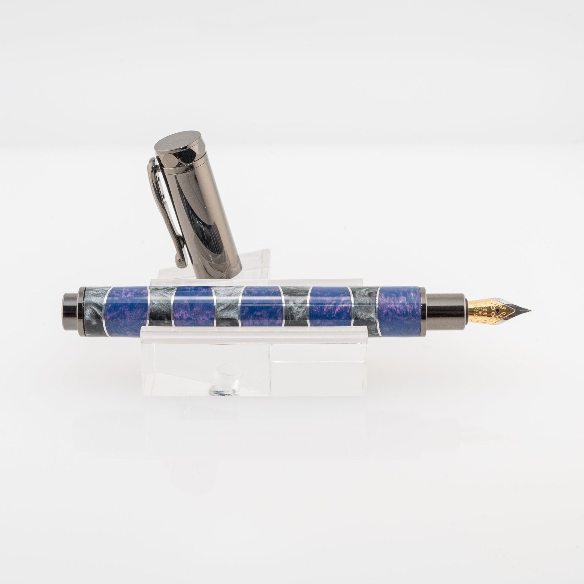 A handmade purple and silver resin and aluminum fountain pen rests on a clear stand. The plating is gunmetal and it features a German Iridium nib.