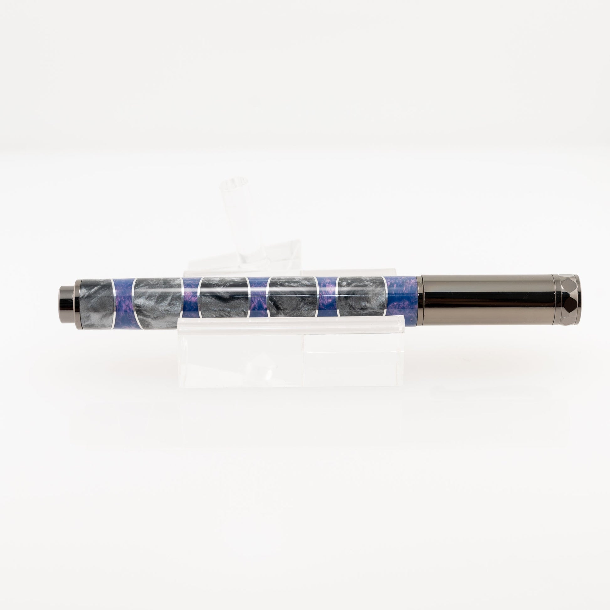 A handmade purple and silver resin and aluminum fountain pen rests on a clear stand. The plating is gunmetal and it features a German Iridium nib.