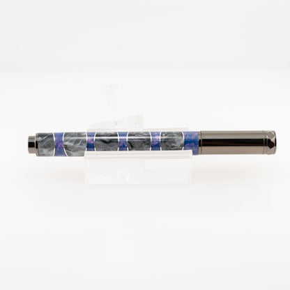 A handmade purple and silver resin and aluminum fountain pen rests on a clear stand. The plating is gunmetal and it features a German Iridium nib.